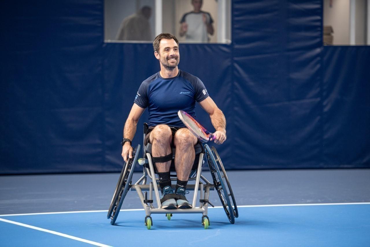 Photo d’Olivier Langlois dans un fauteuil roulant avec une raquette de tennis sur un terrain. 
Crédit : Actu fr
